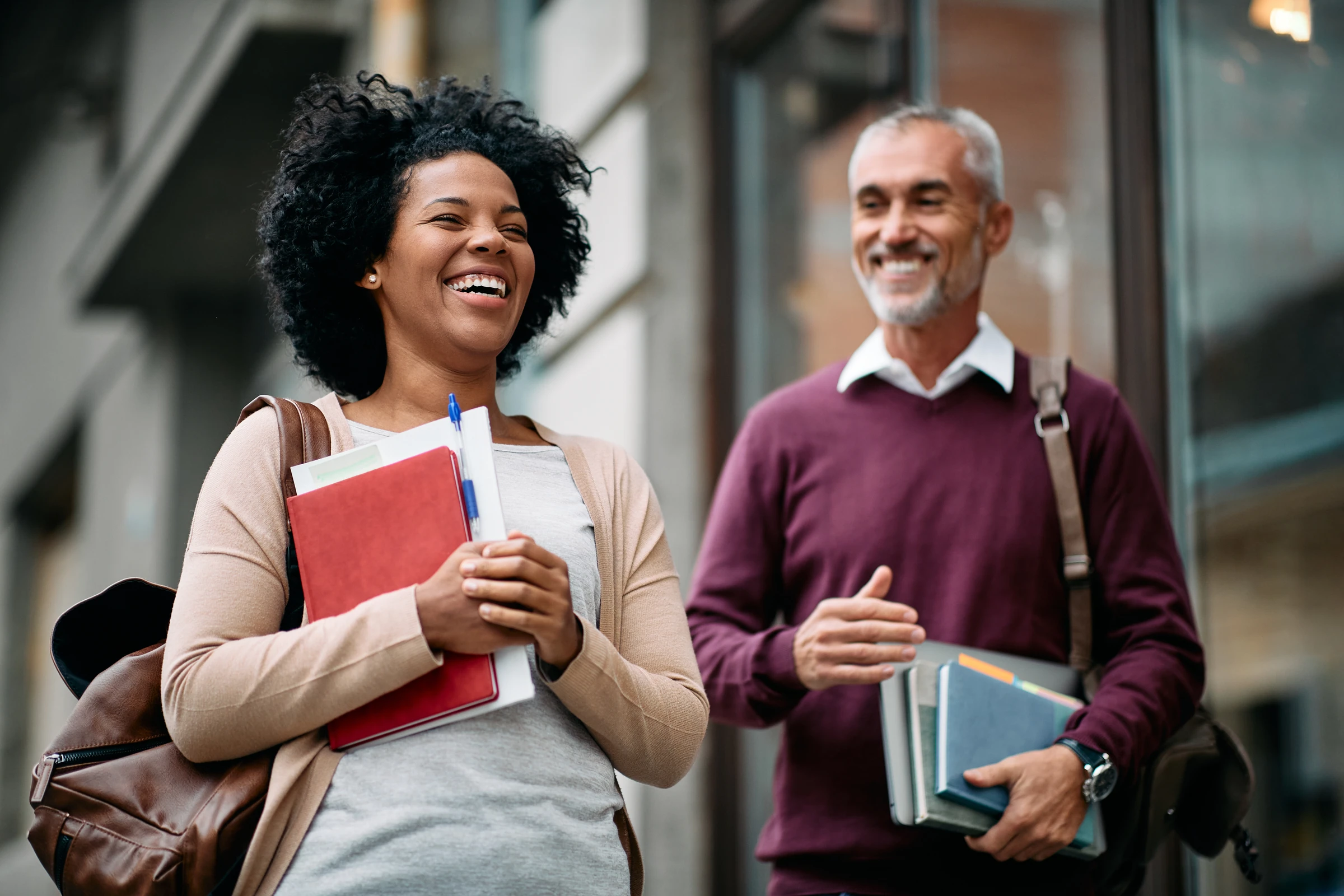 Two adults smiling and carrying books and notebooks outside a building.