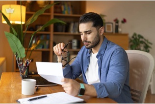 Man reading a letter or document at a home desk, glasses set aside.